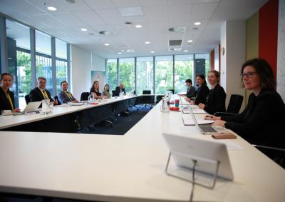 9 people in professional attire seated on both sides of a long table in a sunlit room.