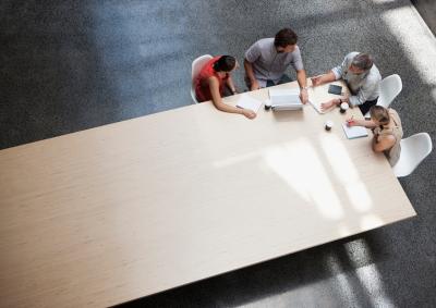 POV from the ceiling down to a long rectangular table with four office workers seated around a corner.